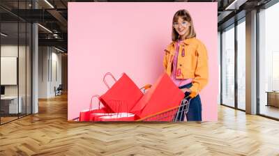 Young woman in sunglasses with price tags on jacket standing near cart with shopping bags on pink background Wall mural