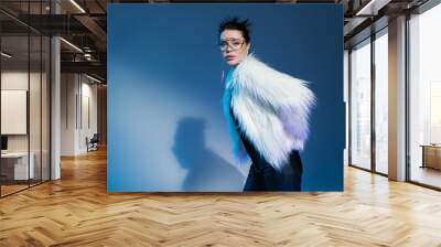 young woman in transparent eyeglasses and fluffy faux fur jacket looking at camera on blue background. Wall mural