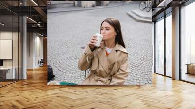 young woman in trench coat drinking coffee to go on cafe terrace. Wall mural