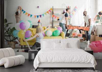 young woman sitting on floor with colorful balloons and her friends decorating room behind Wall mural