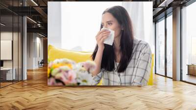 young woman wiping nose while suffering from allergy near flowers on blurred foreground Wall mural