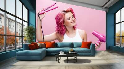 young woman with colorful hair and closed eyes holding straightener and hairdryer isolated on pink Wall mural