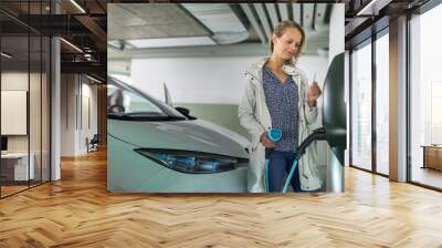 Young woman charging an electric vehicle in an underground garage equiped with e-car charger. Car sharing concept. Wall mural