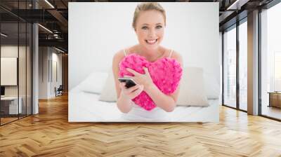 Natural cheerful blonde holding heart pillow and smartphone Wall mural