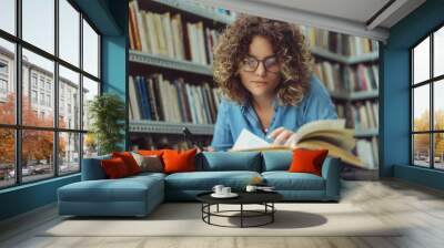 Young female student at the library. Young student reading book at library. Student learning for exam at library. Wall mural