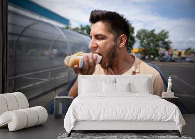 Handsome young brunette man eating hot dog in the parking lot near the gas station. Wall mural