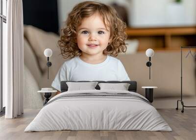 A cheerful child with curly hair stands proudly in a warm, cozy room, showcasing a plain white t-shirt mockup against a backdrop of wooden furniture Wall mural