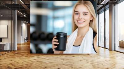 A cheerful woman wearing athletic clothing holds a shaker and has a towel draped around her neck, standing in a bright gym filled with natural light Wall mural