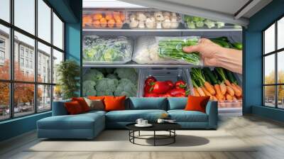 A hand reaches for a bag of green beans in a refrigerator. The refrigerator is stocked with fresh produce, including broccoli, carrots, red peppers, and mushrooms Wall mural