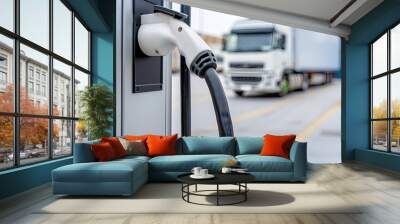 A white electric charging cable is plugged into a charging station. A semi-truck is parked in the background. Wall mural