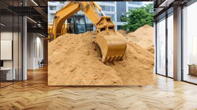 A yellow excavator is actively moving sand and gravel, while workers in safety gear monitor the construction site and surroundings near an industrial building Wall mural