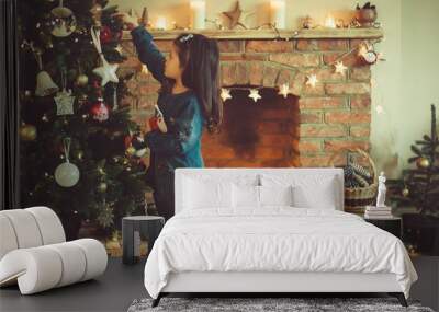 Little girl decorating Christmas tree in a dark room, Christmas setting background; fireplace with lit up star decorations, candles, pine cones in the basket, selective focus, toned photo Wall mural