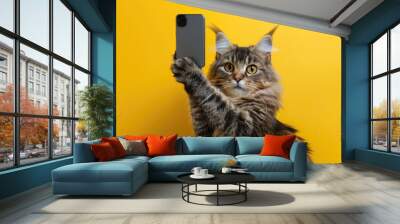 Playful cat taking a selfie on a yellow background, Posing with a smartphone in its paw. Wall mural