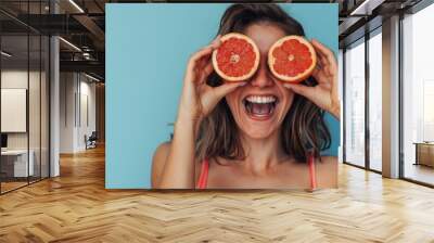 A woman playfully holds two halves of a grapefruit in front of her eyes, smiling brightly against a blue background Wall mural