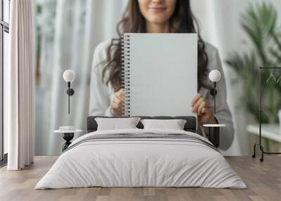 A woman stands in a bright, inviting room, proudly presenting a blank spiral notebook for creative ideas and inspiration Wall mural