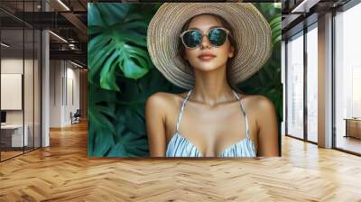 A woman wearing a straw hat and sunglasses is posing in front of a green plant Wall mural