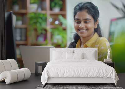 A young Indian businesswoman smiles brightly as she works on her laptop in a modern office setting. She is surrounded by plants and has a professional appearance Wall mural