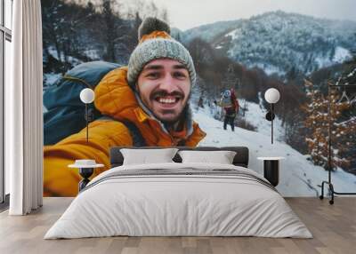 A young man dressed in warm winter clothing is joyfully taking a selfie while hiking on a snowy mountain path. Another hiker is visible in the background, enjoying the scenery Wall mural