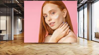 Closeup beauty portrait of redhead caucasian beautiful female applying moisturizing cream or face primer and looking at camera over pink background. Youth and skin care concept. Wall mural