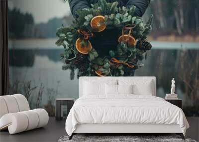 In a peaceful winter landscape, a person stands at the edge of a calm lake, proudly holding an oversized wreath filled with evergreen, oranges, and pinecones, radiating festive spirit Wall mural