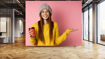 Optimistic happy girl wearing casual clothes holding copy space in one hand, disposable cup of hot drink in other hand, keeping palm open smiling posing on pink background. Wall mural