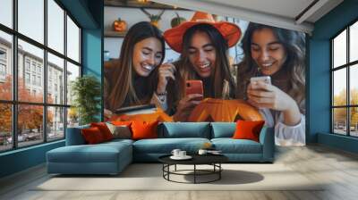 Three friends share laughter and excitement while looking at their phones next to carved jack-o'-lanterns in a festive Halloween atmosphere Wall mural