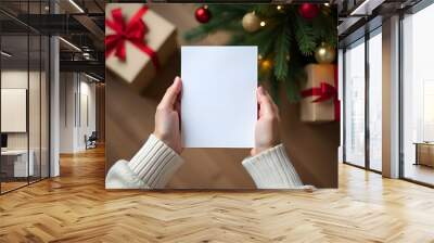 Hands of woman holding a white sheet  paper. Christmas tree and gift beige with red ribbons boxes in the background. Wall mural