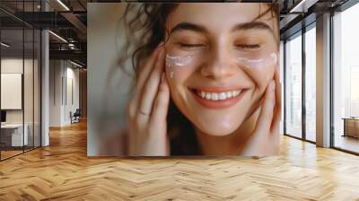 Happy young woman with closed eyes applies cream on her face and moisturizes her skin with cosmetics creating a home spa care concept Wall mural