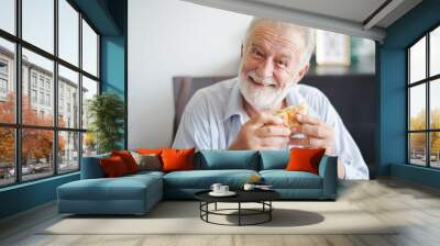 Seniors European man is sitting to eat a burger at home which looking at camera. Retired man is smiling with felling happy and take a burger on hand. Wall mural