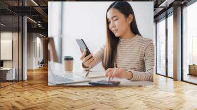 Young woman using a calculator and other hand using phone to chat with customer and have notebook on the table at cafe or co working space which smiling and felling happy. co working space concept. Wall mural