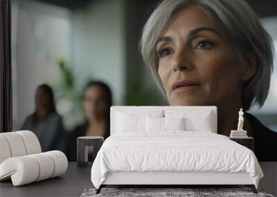 Close-up of senior businesswoman in an office meeting room, blurred coworkers in the background, minimal background focusing on her expression. Wall mural