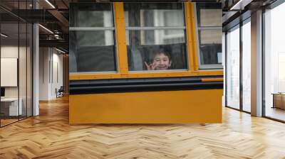 Back to School lifestyle portrait of young boy on yellow school bus smiling looking out the window and showing the sign for I-Love_you with his hand  Wall mural