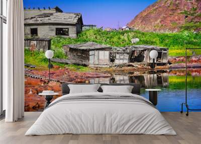 Old ruined wooden houses reflected in bay water in Teriberka, Murmansk oblast, Kola peninsula, Russia. Beautiful north nature, red rocks and dilapidated dwelling abandoned by man Wall mural