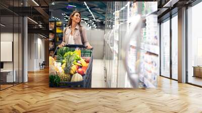 Beautiful female with shopping cart walking by supermarket freezer choosing what to buy. Buying food in grocery store. Wall mural