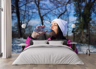 Female athlete in winter clothes drinking water before training on snow. Jogging and recreation. Wall mural