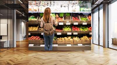 Good looking woman standing in front of vegetable shelves choosing what to buy. Buying groceries and healthy organic food in supermarket. Wall mural