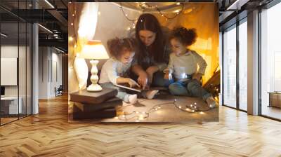 Reading and family games in children's tent. Mother and two twins daughters with books and flashlight before going to bed. Wall mural