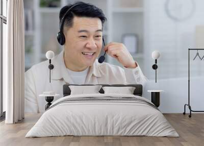 A cheerful Asian man wearing a headset smiles while working from his home office. He is interacting and providing customer support with positivity and professionalism. Wall mural