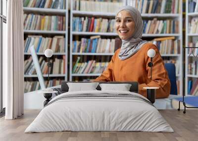 A cheerful young woman wearing a hijab sits at a library table, using her laptop among rows of books. Her smile indicates confidence and contentment with her study environment. Wall mural