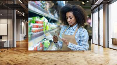 A grocery store employee in an apron checks the inventory on the shelves using a tablet. The worker appears focused and attentive, ensuring the store is well-stocked and organized for customers. Wall mural