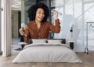 A joyful African American businesswoman celebrates a successful transaction or achievement at her modern office desk, holding a credit card and showing a victory gesture. Wall mural