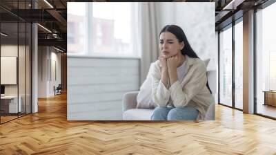 A sad and worried young woman suffers from depression, mental disorders. She sits thoughtfully in the hospital room on the sofa by the window, resting her head on her hands. Wall mural