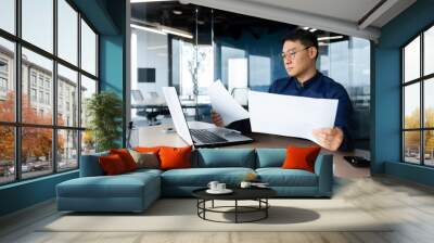 A serious young man, an Asian businessman, works in the office at the table, holds and looks at documents, graphs, plans, bills. Wall mural