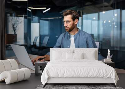 A serious young man accountant, financier, analyst, auditor sits in the office at the table. He holds documents and a pen in his hands, checks accounts, finances, types on a laptop. Wall mural