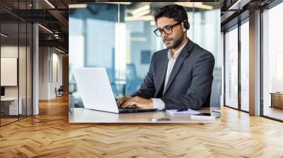 A young Indian businessman is working in the office on a laptop and wearing headphones. Concentrated typing on the keyboard, chats. Wall mural