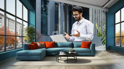 A young Indian male office worker is having a video call, sitting on a bench near a building and holding a laptop on his lap. Wall mural