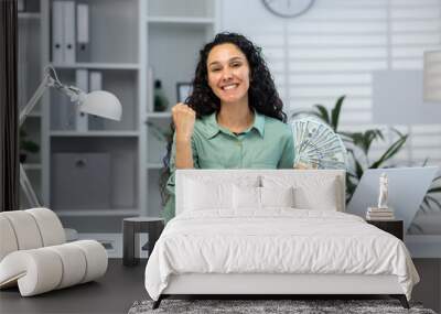 A young Latin American woman is sitting at the desk in the office and holding cash money in her hands. Rejoices and smiles at the camera, shows a victory hand gesture yes. Wall mural