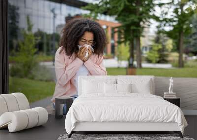 African American woman sneezing into a tissue while sitting outside. She appears to be experiencing allergy or cold symptoms. The surroundings suggest it is a park or urban area, with green trees Wall mural