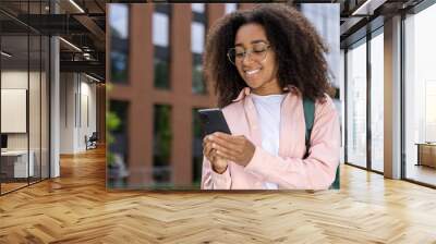 African American woman student with glasses and backpack smiling while using smartphone outdoors in urban setting. Confident, connected, and engaged in a city environment. Wall mural