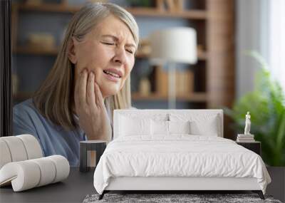 An elderly Caucasian woman sits at home, touching her cheek and grimacing in pain. She appears to be suffering from a severe toothache or jaw discomfort. Wall mural